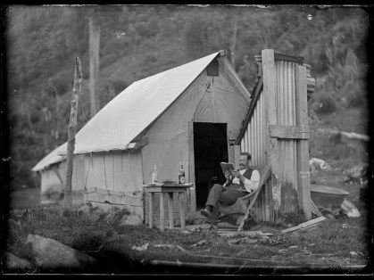 full_A_man__outside_a_hut__sitting_in_a_deck_chair__reading_a_book.